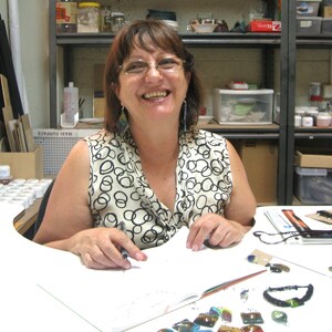 Pode incluir: Uma mulher de &oacute;culos e top estampado sorri sentada a uma mesa. Ela segura uma caneta e olha para um caderno. V&aacute;rios materiais de arte e pe&ccedil;as acabadas est&atilde;o sobre a mesa.