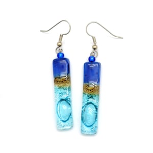 Skinny rectangle glass blue earrings. The very top has a cobalt blue glass bead. The fused glass is in three bands of color. At the top is dark cobalt blue. Second, bronze. Third and widest is an aqua turquoise color. The glass has bubbles.