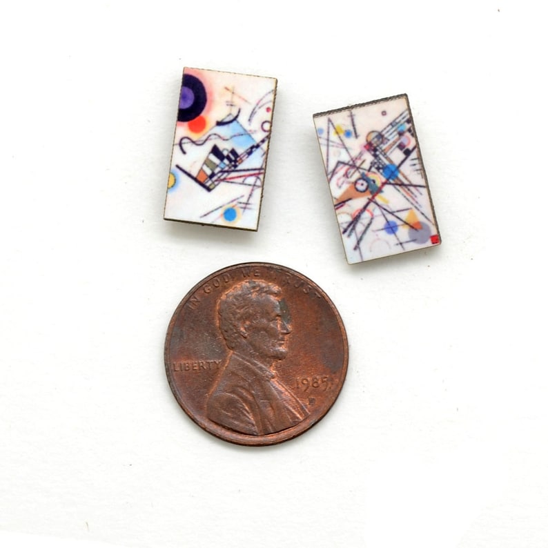 Small rectangle stud earrings featuring Kandinsky geometric art in a rainbow of circles, lines and squiggles. The earrings are next to a copper USA penny showing the earrings are smaller than the penny.