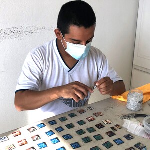 A male artist is wearing a Covid protection mask and making jewelry at a desk.