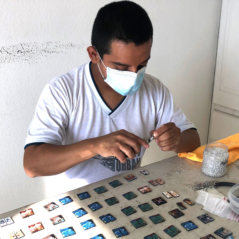 A male artist is wearing a Covid protection mask and making jewelry at a desk.