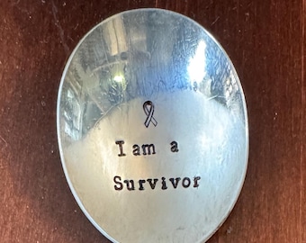 May include: A silver spoon with the words "I am a Survivor" stamped on the bowl. A purple ribbon is above the text. The handle has a decorative floral design.