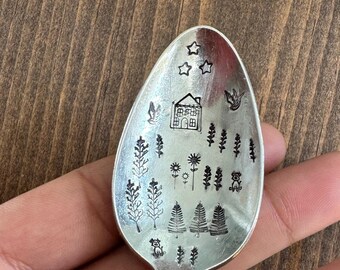 May include: A silver spoon with a nature-themed design. The spoon's bowl features etched images of a house, trees, stars, and other natural elements. The spoon is held against a wooden background.