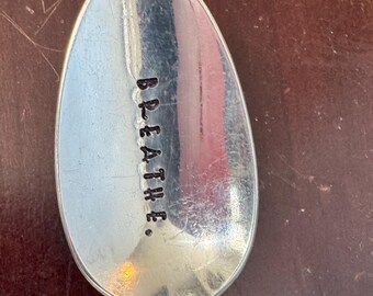 May include: A silver spoon with the word "BREATHE." stamped in black letters on the spoon's bowl. The spoon is set against a dark brown surface.