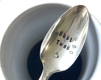 May include: A silver teaspoon with the words "Best Teas" engraved on it, along with a small teacup and two hearts. The spoon is resting in a white teacup with dark liquid inside.