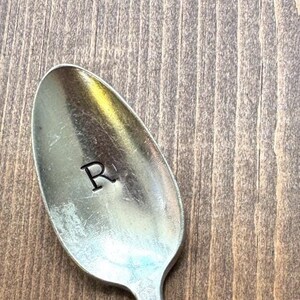May include: A silver spoon with an ornate design on the handle. The spoon has the letter "R" engraved on the bowl.