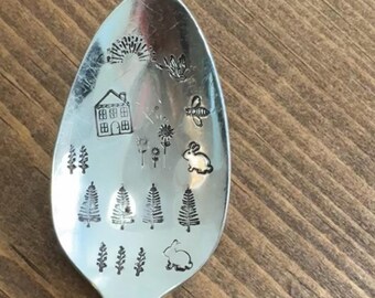 May include: A silver spoon with etched designs of a house, trees, flowers, a bunny, and a sun. The spoon is set against a wooden background. The spoon's handle has vertical lines.