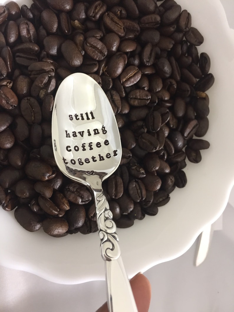 May include: A silver spoon with the words "still having coffee together" engraved on it, resting on a bed of coffee beans.