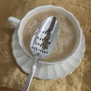 May include: A silver spoon with the words "still having coffee together" engraved on it. The spoon is resting in a white coffee cup with coffee inside.