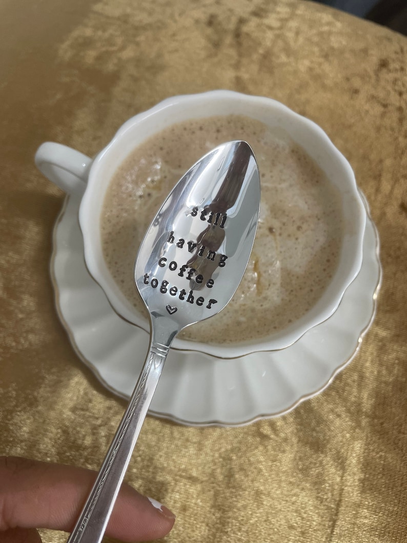 May include: A silver spoon with the words "still having coffee together" engraved on it. The spoon is resting in a white coffee cup with coffee inside.