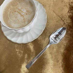 May include: A silver spoon with the inscription "still having coffee together" on a gold velvet surface. A white cup with coffee is in the background.