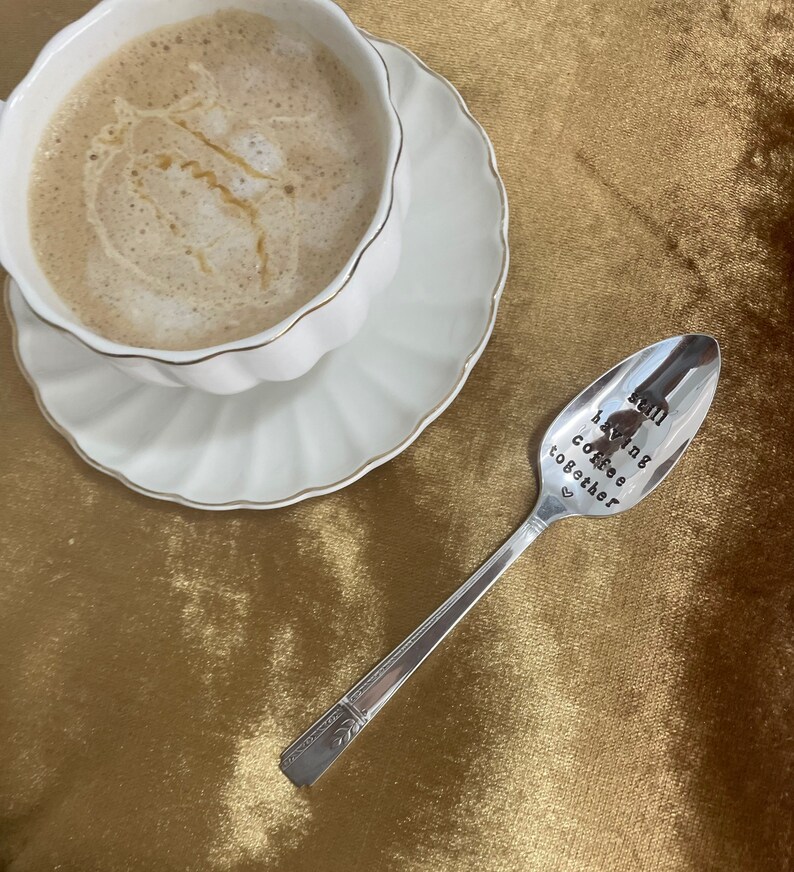 May include: A silver spoon with the inscription "still having coffee together" on a gold velvet surface. A white cup with coffee is in the background.