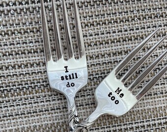May include: Two silver forks with the words "I still do" and "Me too" engraved on them. The forks are on a silver and white patterned surface.