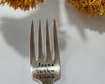 May include: A silver fork with the stamped text "Jesus is the Lamb." The fork has four tines and a smooth handle. Dried orange flowers are in the background.