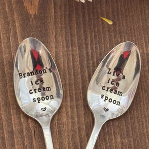 May include: Two silver ice cream spoons with stamped text. One spoon reads "Brandon's ice cream spoon" and the other reads "Liv's ice cream spoon". Each spoon has a small heart symbol. The spoons are on a brown wooden surface.
