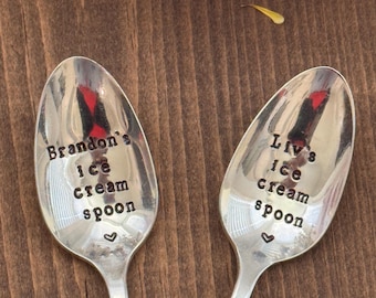 May include: Two silver ice cream spoons with stamped text. One spoon reads "Brandon's ice cream spoon" and the other reads "Liv's ice cream spoon". Each spoon has a small heart symbol. The spoons are on a brown wooden surface.