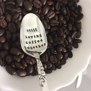 May include: A silver spoon with a heart and the words "still having coffee together" engraved on the bowl. The spoon is surrounded by coffee beans in a white bowl.
