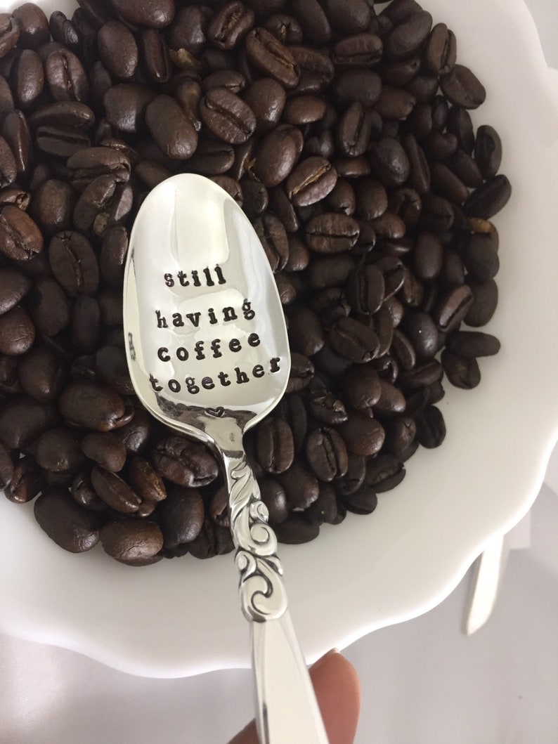 May include: A silver spoon with a heart and the words "still having coffee together" engraved on the bowl. The spoon is surrounded by coffee beans in a white bowl.