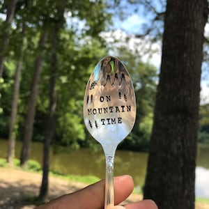 May include: A silver spoon with the words "On Mountain Time" engraved on it. The spoon is held in a hand against a blurred background of trees and a lake.