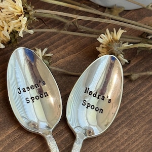 May include: Two silver spoons with engraved text. One spoon reads "Jasen's Spoon" and the other reads "Nedra's Spoon". The spoons are set on a wooden surface, with dried flowers in the background.