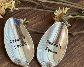 May include: Two silver spoons with engraved text. One spoon reads "Jasen's Spoon" and the other reads "Nedra's Spoon". The spoons are set on a wooden surface, with dried flowers in the background.