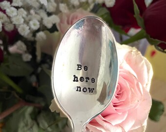 May include: A silver spoon with the stamped words "Be here now." The spoon is in front of a blurred background of pink and red roses and white flowers. The spoon has a reflective surface.