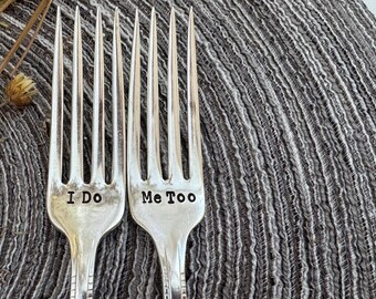 May include: Two silver forks with the words "I Do" and "Me Too" stamped on them. The forks are set against a textured gray background, suggesting a wedding or celebratory theme.