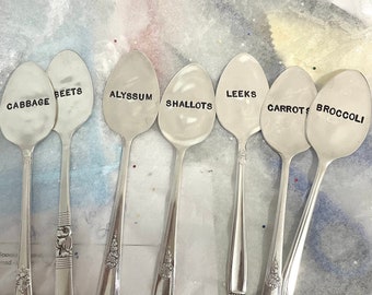 May include: Seven silver spoons with black lettering on the bowl of each spoon. The spoons are labeled "Cabbage", "Beets", "Alyssum", "Shallots", "Leeks", "Carrots", and "Broccoli".