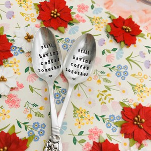 May include: Two silver spoons with the words "still having coffee together" engraved on them. The spoons are on a white background with a floral pattern.