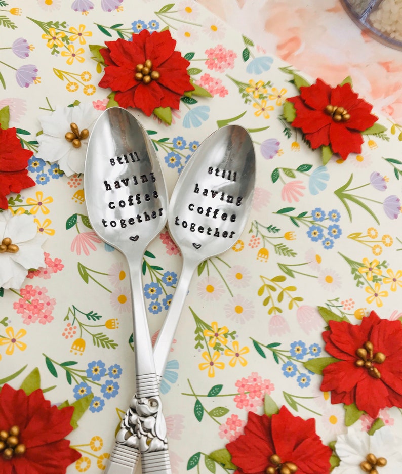 May include: Two silver spoons with the words "still having coffee together" engraved on them. The spoons are on a white background with a floral pattern.