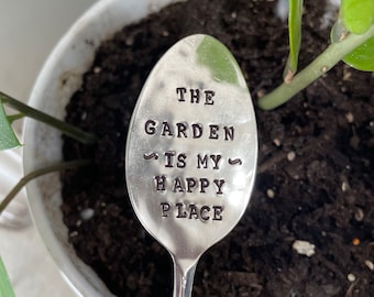 May include: A silver spoon with the words "The Garden is my Happy Place" engraved on it. The spoon is sitting in a pot of soil with a green plant.