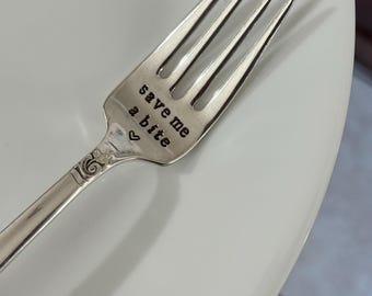 May include: A silver fork with four tines, resting on a white plate. The fork's handle is engraved with the words "save me a bite" and a small heart symbol. The fork has a decorative floral design.