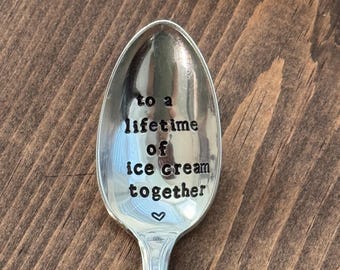 May include: A silver spoon with the words "to a lifetime of ice cream together" stamped on the bowl, with a small heart symbol below the text. The spoon is set against a dark wood grain background.