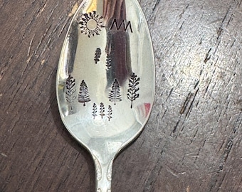 May include: Silver spoon with a nature-themed design. The spoon's bowl features etched images of a sun, mountains, and trees. The handle has an ornate, vintage-style pattern. The spoon is likely made of stainless steel or a similar metal.