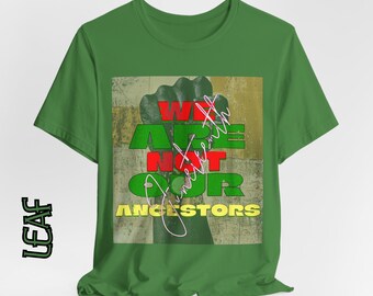 Può includere: T-shirt verde con un design grafico raffigurante un pugno alzato e la scritta "WE ARE NOT OUR ANCESTORS". Il testo è in rosso, verde e giallo. La maglietta ha un girocollo e maniche corte.