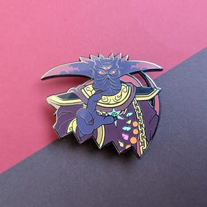 May include: A purple and gold enamel pin depicting a three-eyed creature wearing a crown and a long, flowing robe. The creature is holding a green gem in its hand.