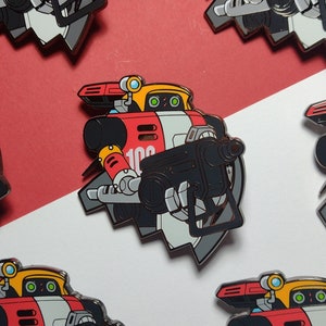 May include: Enamel pin featuring a red, yellow, and gray robot holding a black weapon. The robot has the number "100" on its chest. The background is red and white.
