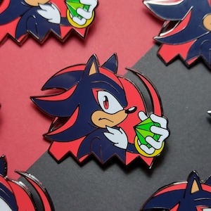 May include: A metal enamel pin featuring the character Shadow the Hedgehog in a dark blue and black color scheme with red accents. The character is depicted holding a bright green faceted gemstone in one hand, set against a red circular background.