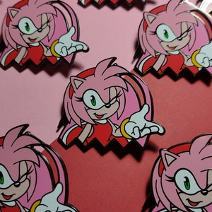 May include: Enamel pins featuring Amy Rose from the Sonic the Hedgehog series. The pins depict Amy with pink hair, a red dress, and white gloves, set against a pink and red background. The pins have a black outline and a gold bracelet.