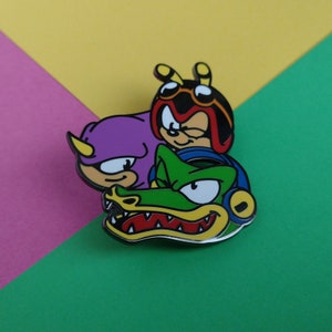 May include: A green crocodile, a purple character with a yellow horn, and a red and black character with a yellow bee antenna and goggles are stacked on top of each other. The characters are enamel pins.