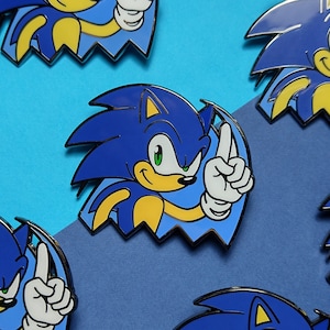 May include: A collection of enamel pins featuring the character Sonic the Hedgehog. Each pin displays the blue character with yellow accents, green eyes, and white gloves, shown in a dynamic pose with one finger raised against a jagged blue background.