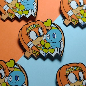 May include: A set of five enamel pins featuring a cartoon character with orange fur and a blue character. The orange character is wearing a gold headband and a yellow heart. The blue character is wearing a yellow heart. The pins are in a ghost shape.