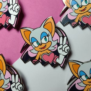 May include: Enamel pins featuring a cartoon bat character with gray fur, orange ears, and blue eyes. The character is giving a peace sign with a white hand. The background is pink and white.