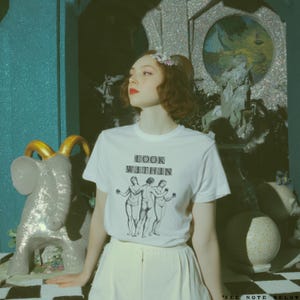 May include: White t-shirt with the text "LOOK WITHIN" above a graphic of three figures. The person is wearing a white skirt and has red lipstick. A glittery elephant statue with gold horns is on the left.