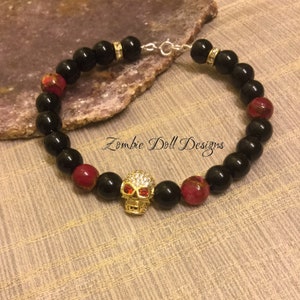 May include: Black and red beaded bracelet with a gold skull charm. The skull has red eyes and is encrusted with rhinestones. The bracelet has a silver clasp.