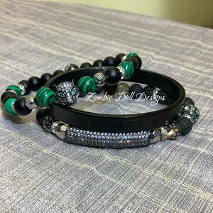Può includere: Un set di quattro braccialetti con perline di onice nero, pietre di malachite verde, un ciondolo di pantera nera e una fascia nera con strass. I braccialetti sono impilati su una superficie di tessuto grigio.