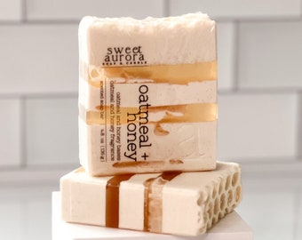 May include: Three bars of oatmeal and honey soap stacked on top of each other. The top bar has the text "sweet aurora soap & candle" and "oatmeal + honey" printed on it. The soap bars are white with a honey-colored swirl.