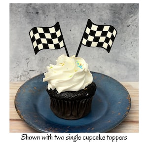 May include: A chocolate cupcake topped with white frosting and two black and white checkered racing flags. The cupcake sits on a blue plate. Text at the bottom reads: Shown with two single cupcake toppers.