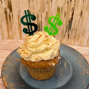 May include: A cupcake with white frosting and a sprinkle of cinnamon sits on a blue plate. Two dollar sign toppers, one green and one dark green, are inserted into the frosting. The cupcake is a dessert item.