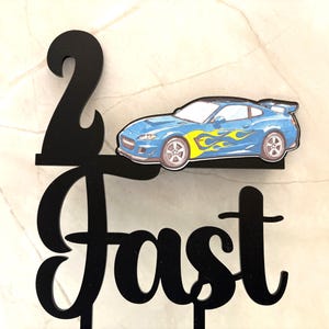 May include: Black cake topper with the number 2 and the word "Fast". A blue sports car with yellow flames is attached to the topper. The cake topper is designed for a birthday celebration.